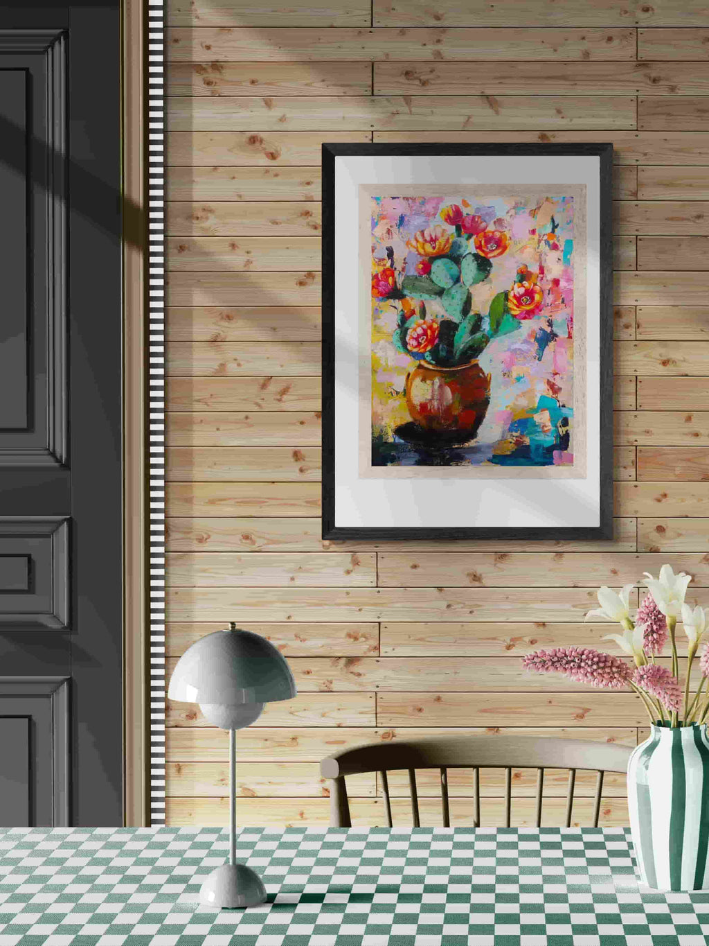 Framed colorful artwork of cacti on a wooden wall above a checkered table.