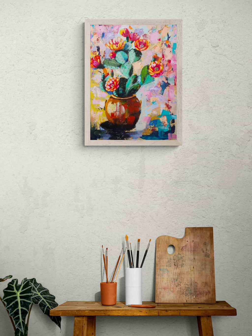 Framed colorful painting of flowers on a wall above a wooden table with art supplies.