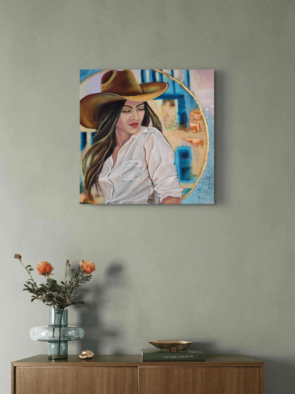 Mixed Media painting of a woman wearing a cowboy hat on a wall above a wooden side table with a vase and decorative items.