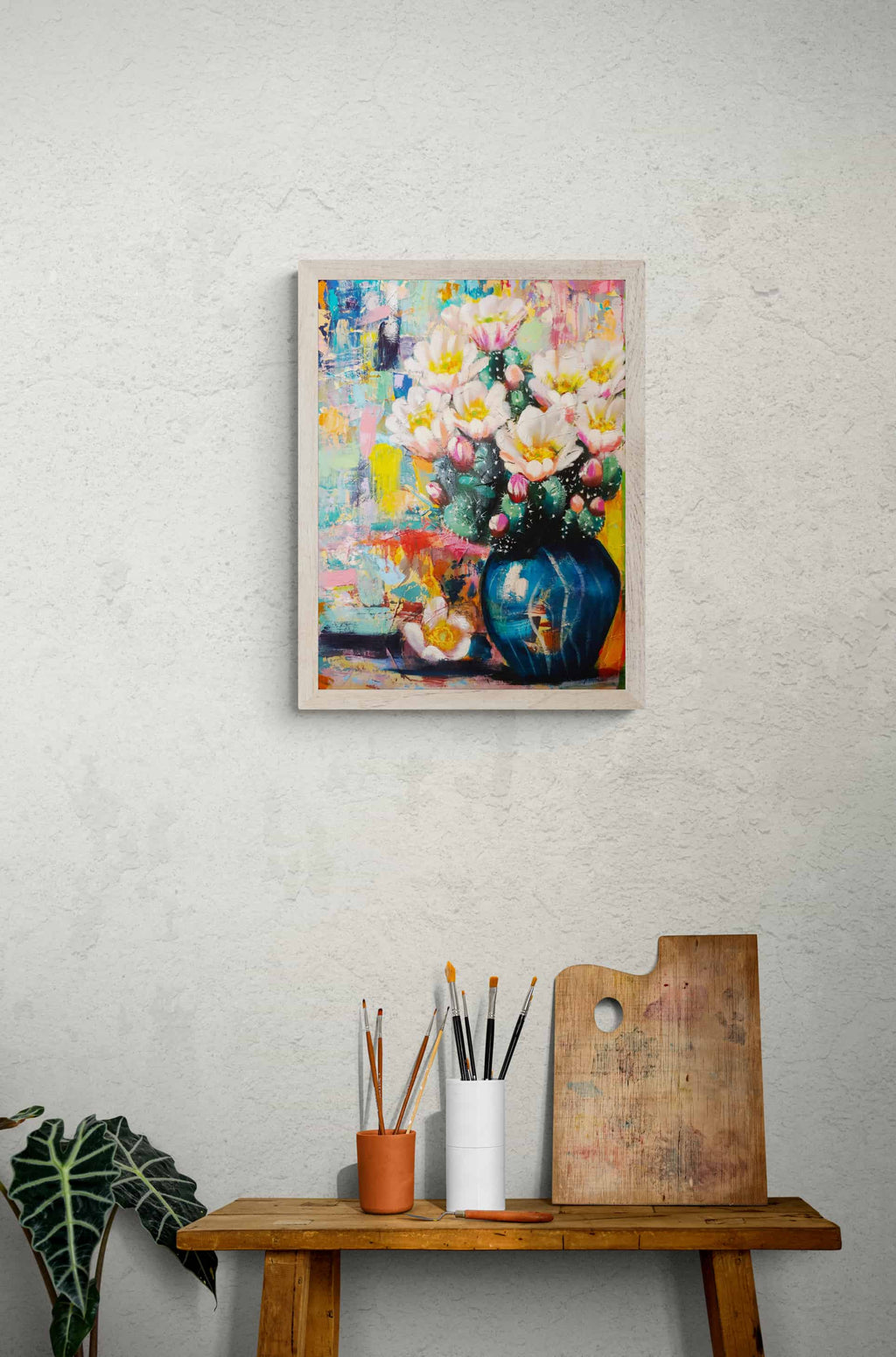 Framed colorful floral painting on a wall above a wooden table with art supplies.