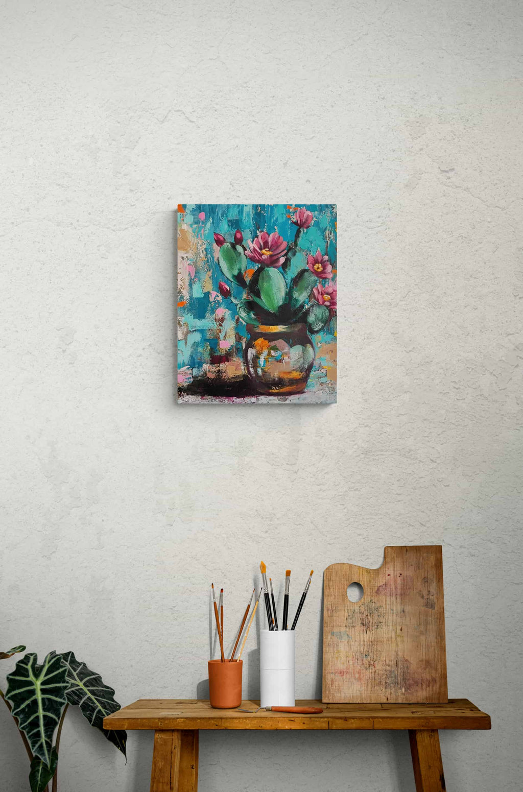 Colorful cactus painting on a textured wall above a wooden table with art supplies.