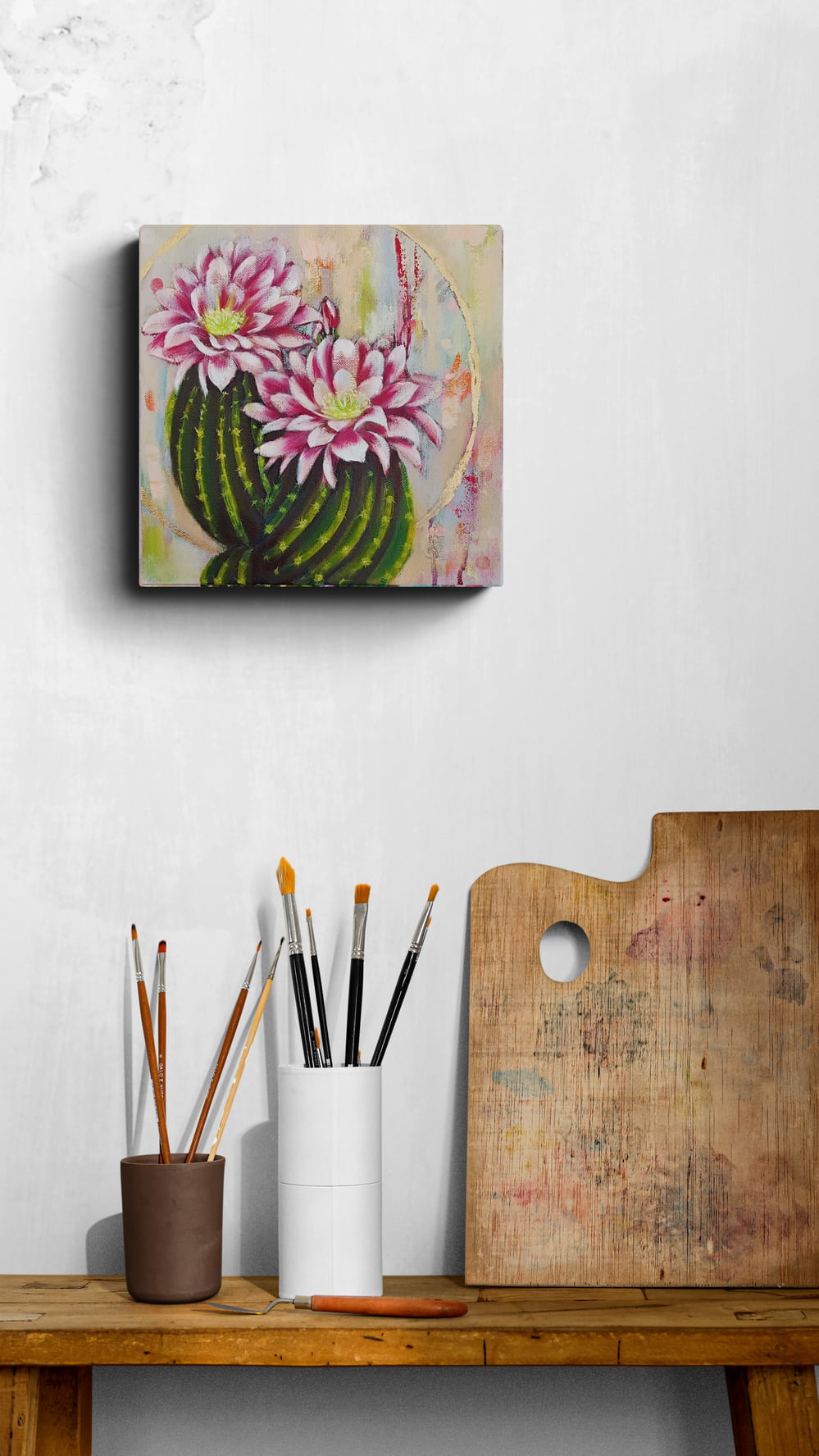 Canvas painting of a cactus with pink flowers on a wall above a wooden table with art supplies.