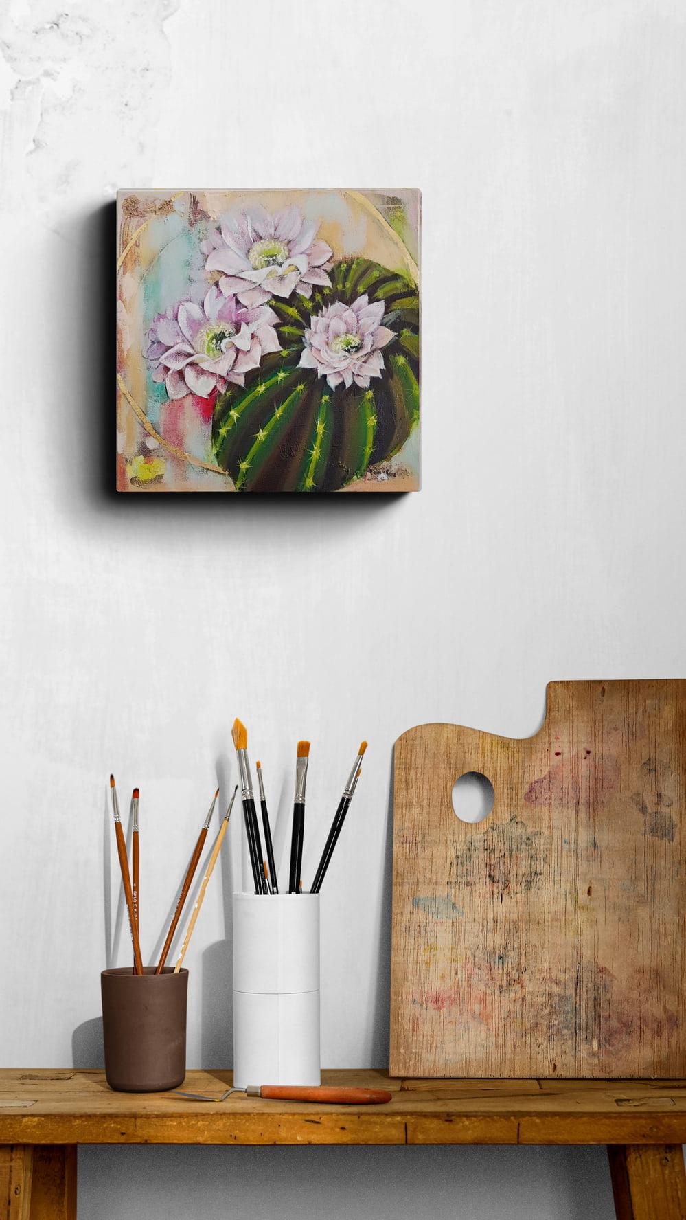 Canvas of a floral cactus painting on a wall above a wooden table with art supplies.