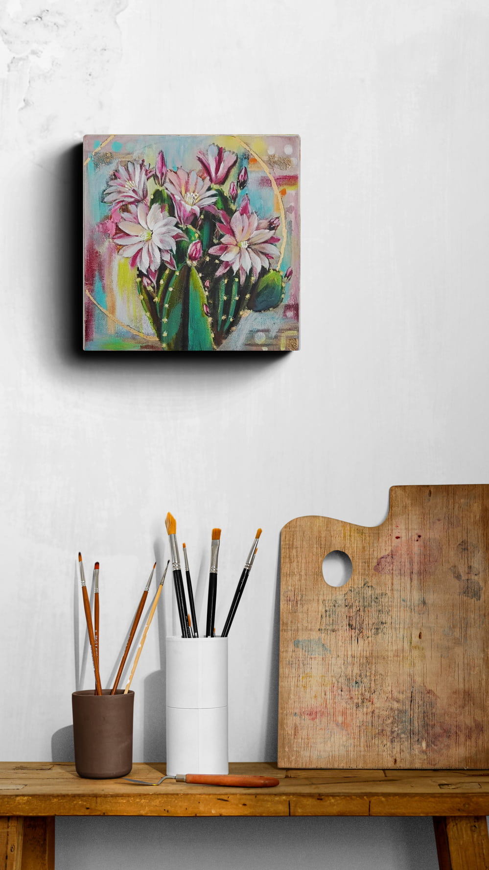 Canvas Painting of a blooming cactus painting on a wall above a wooden shelf with art supplies.