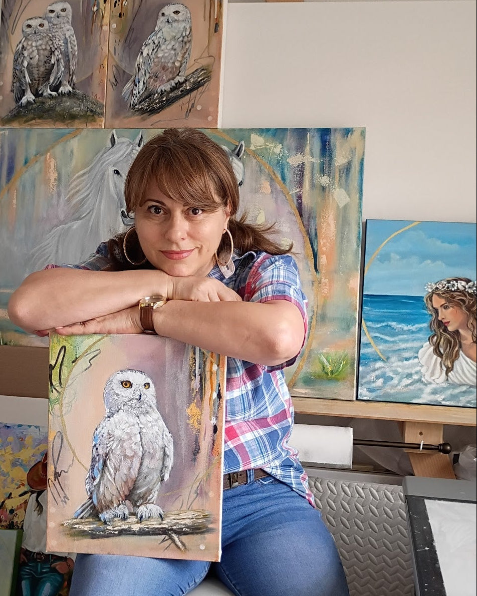 Demy Karalis sitting in her studio. Holding a painting of an owl. Behind her is another paintings of owls, woman and horses.