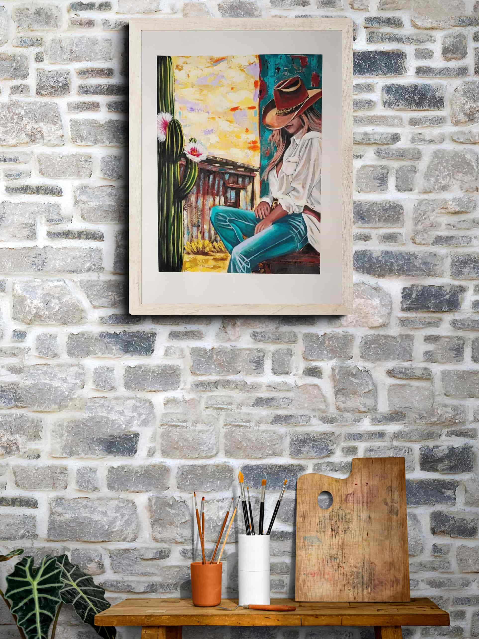 Framed artwork of a person in a cowboy hat on a stone wall with art supplies below.