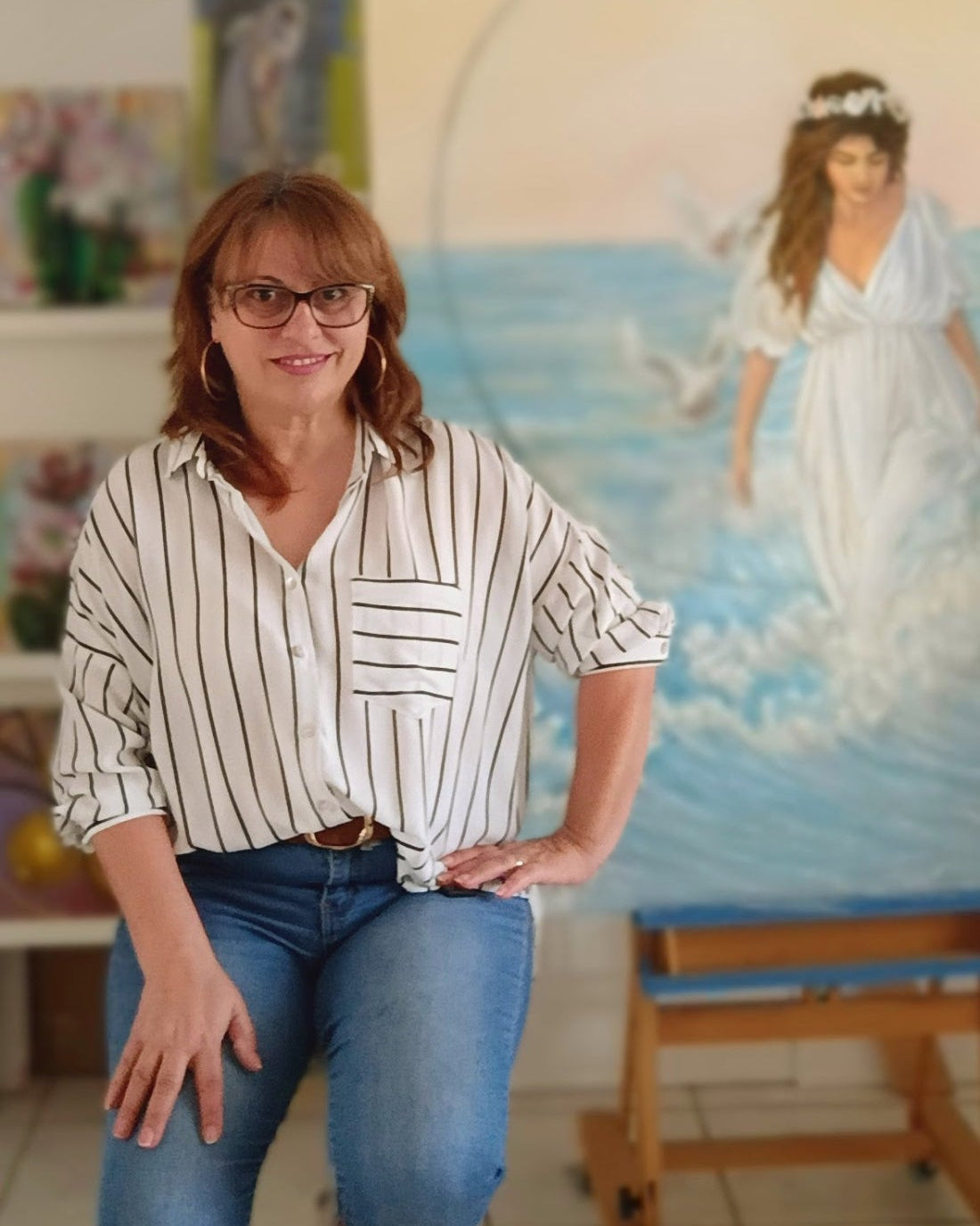 Demy Karalis standing in her studio with a painting of a woman in a white dress on the wall.