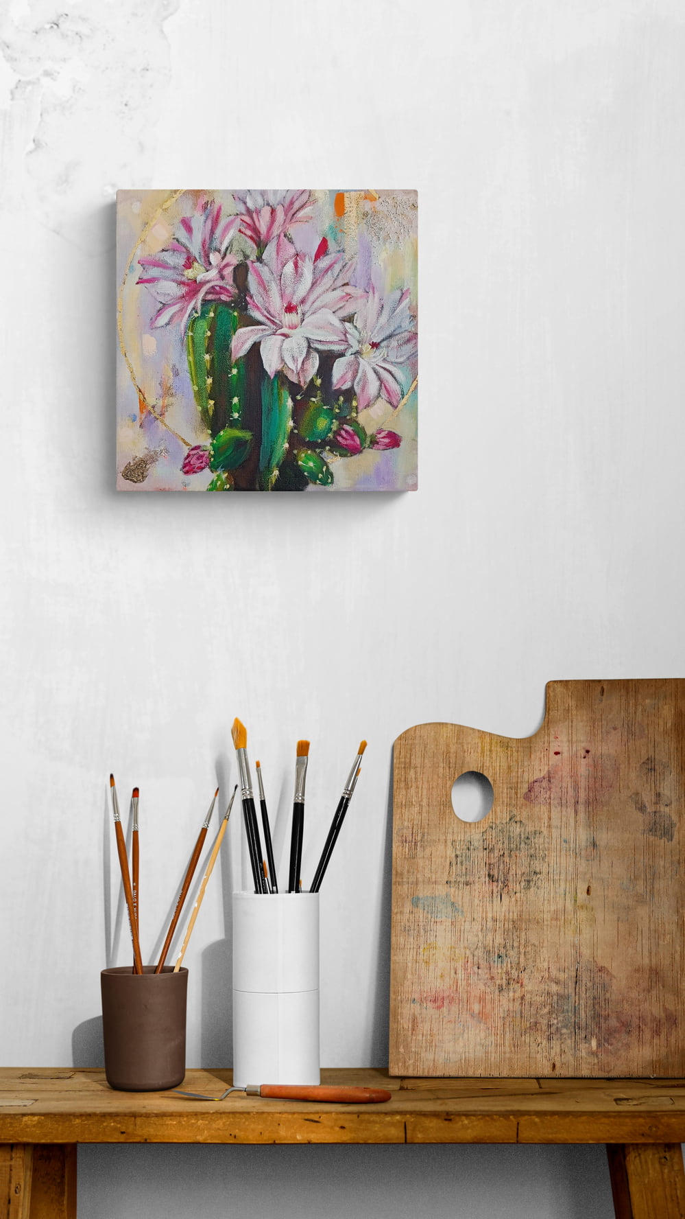 Canvas painting of flowers and cactus on a wall above a wooden table with art supplies.