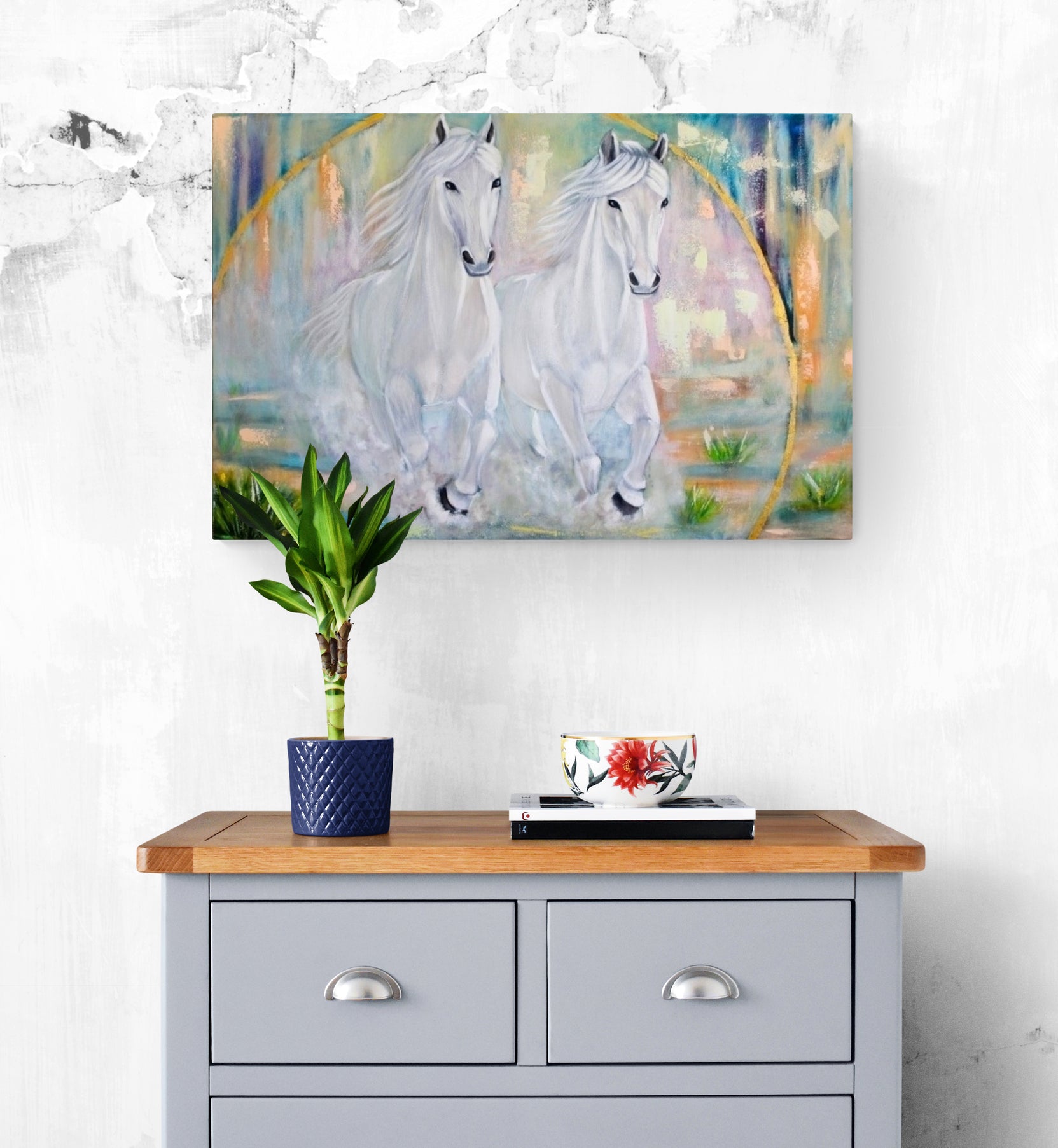 Painting of two white horses on a wall above a wooden table with a plant and decorative items.