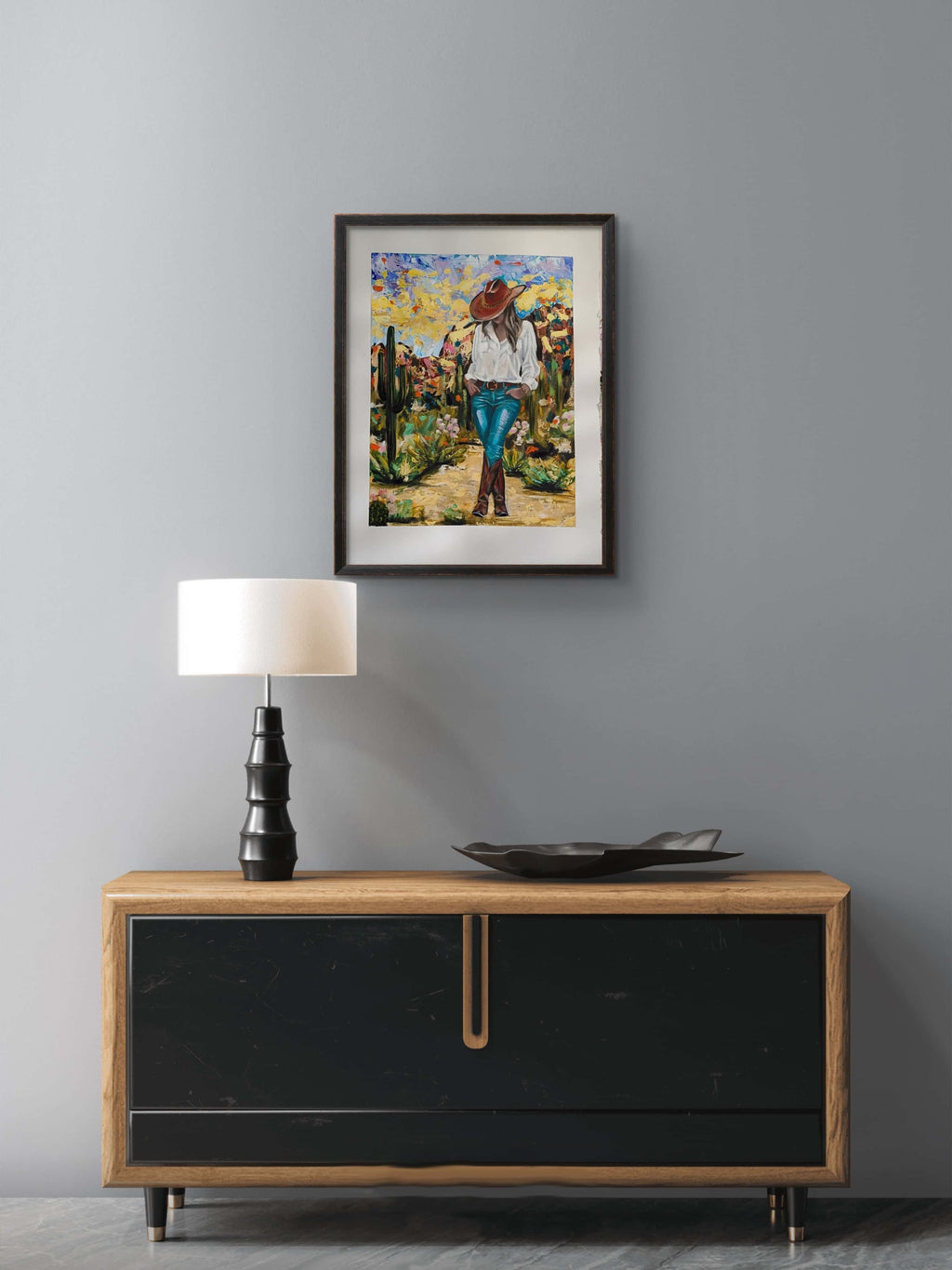 Framed artwork on a gray wall above a wooden sideboard with a lamp and decorative bowl.