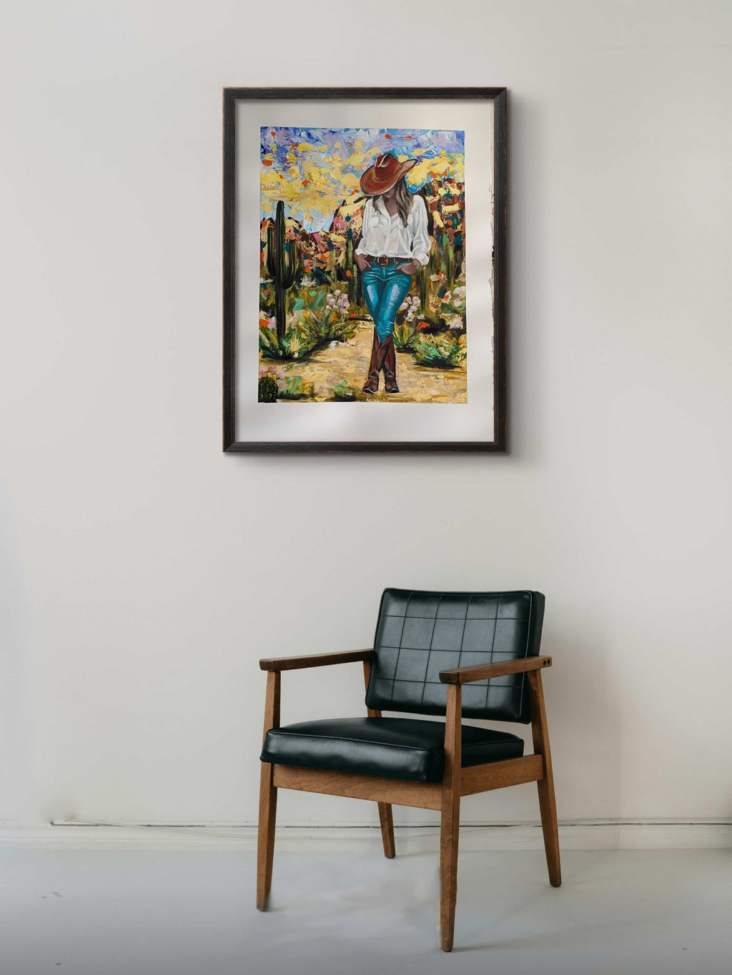 Framed painting of a person in a desert landscape above a black chair.