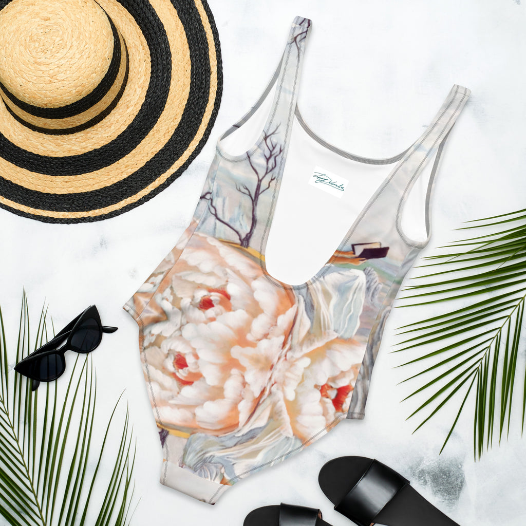 An one piece swimsuit with printed art work on it next to a straw  hat, black sunglasses and  black flip flops. 