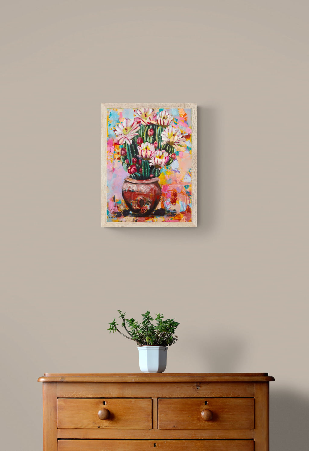 Colorful painting of a cactus with flowers on a beige wall above a wooden dresser.