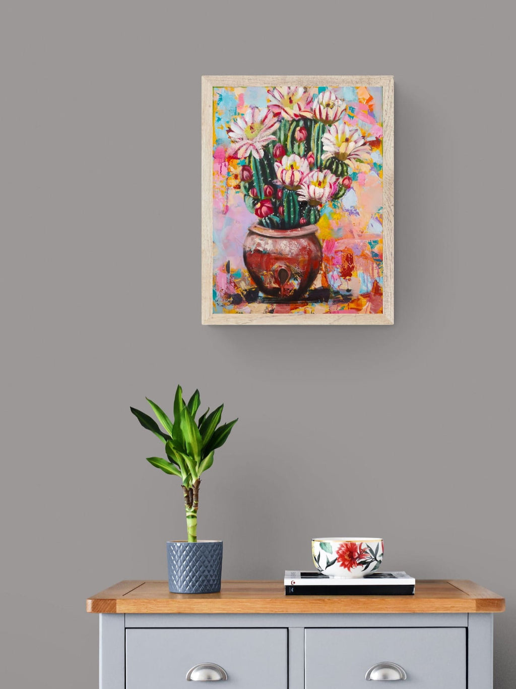 Colorful abstract painting of a cactus with flowers on a gray wall above a wooden cabinet with a plant and decorative items.